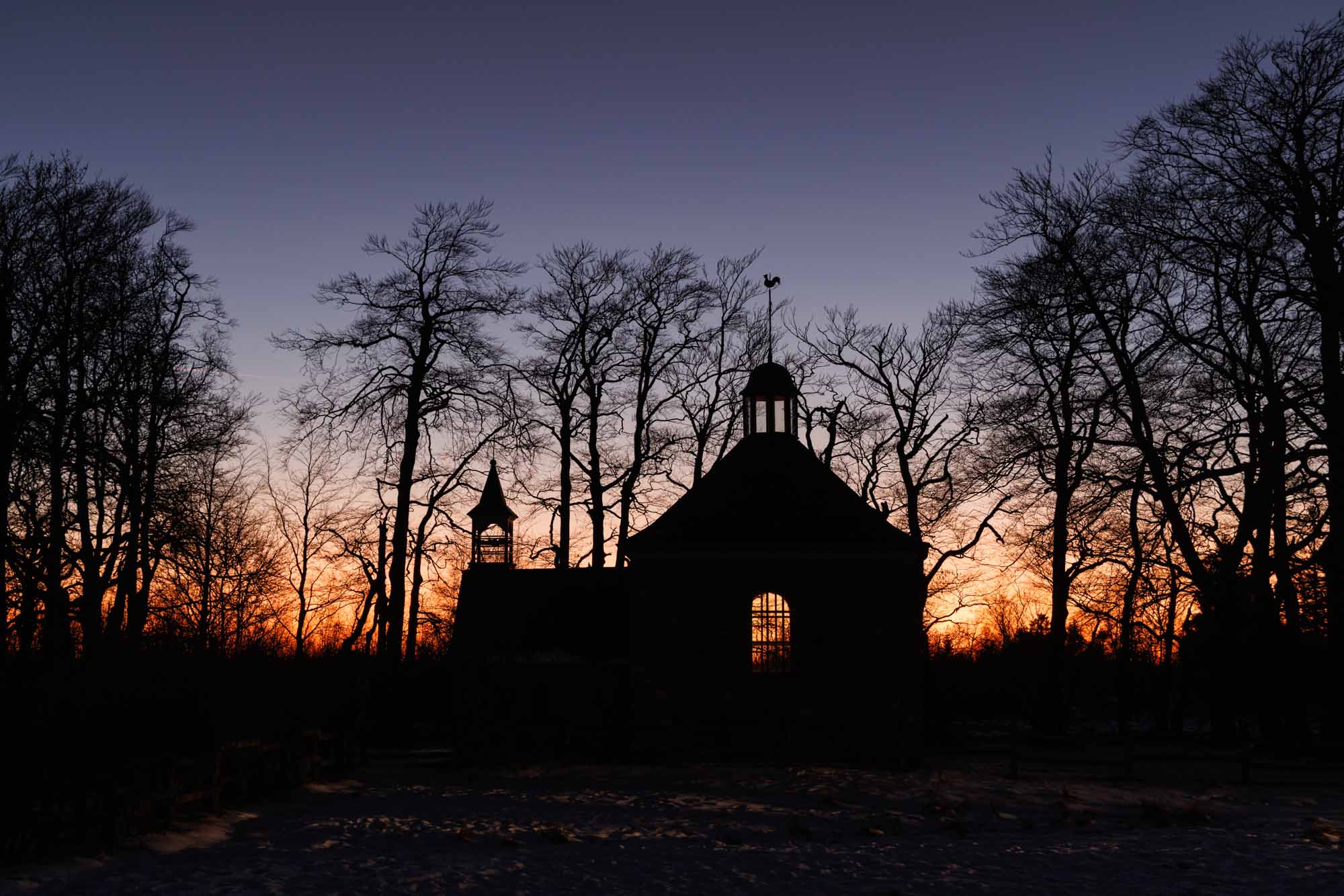 Silhouette de la chapelle Fischbach au coucher du soleil, entourée d’arbres dénudés en hiver.