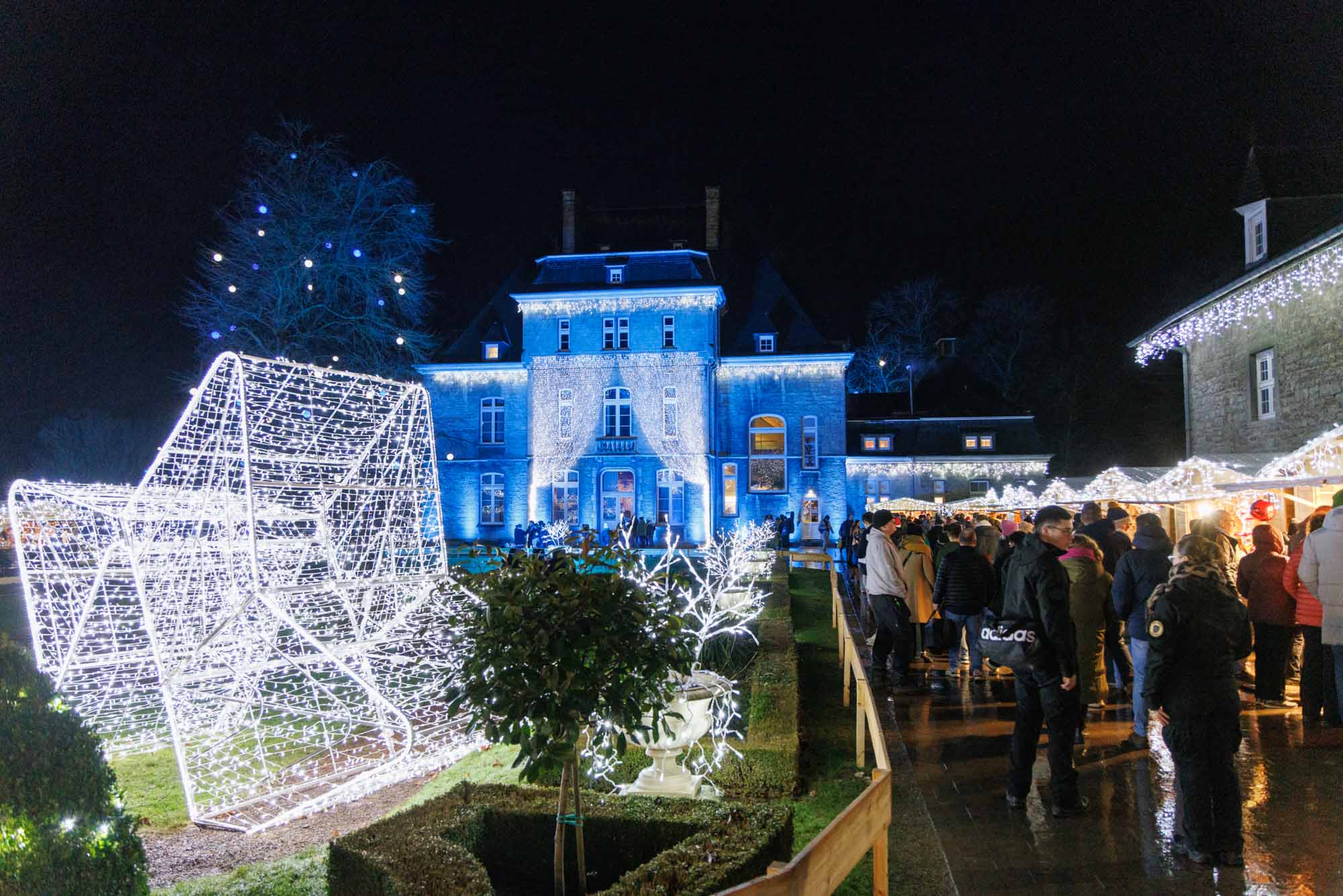 Kerstmarkt van Ciney bij nacht met lichtdecoraties en bezoekers voor een blauw verlicht historisch gebouw.