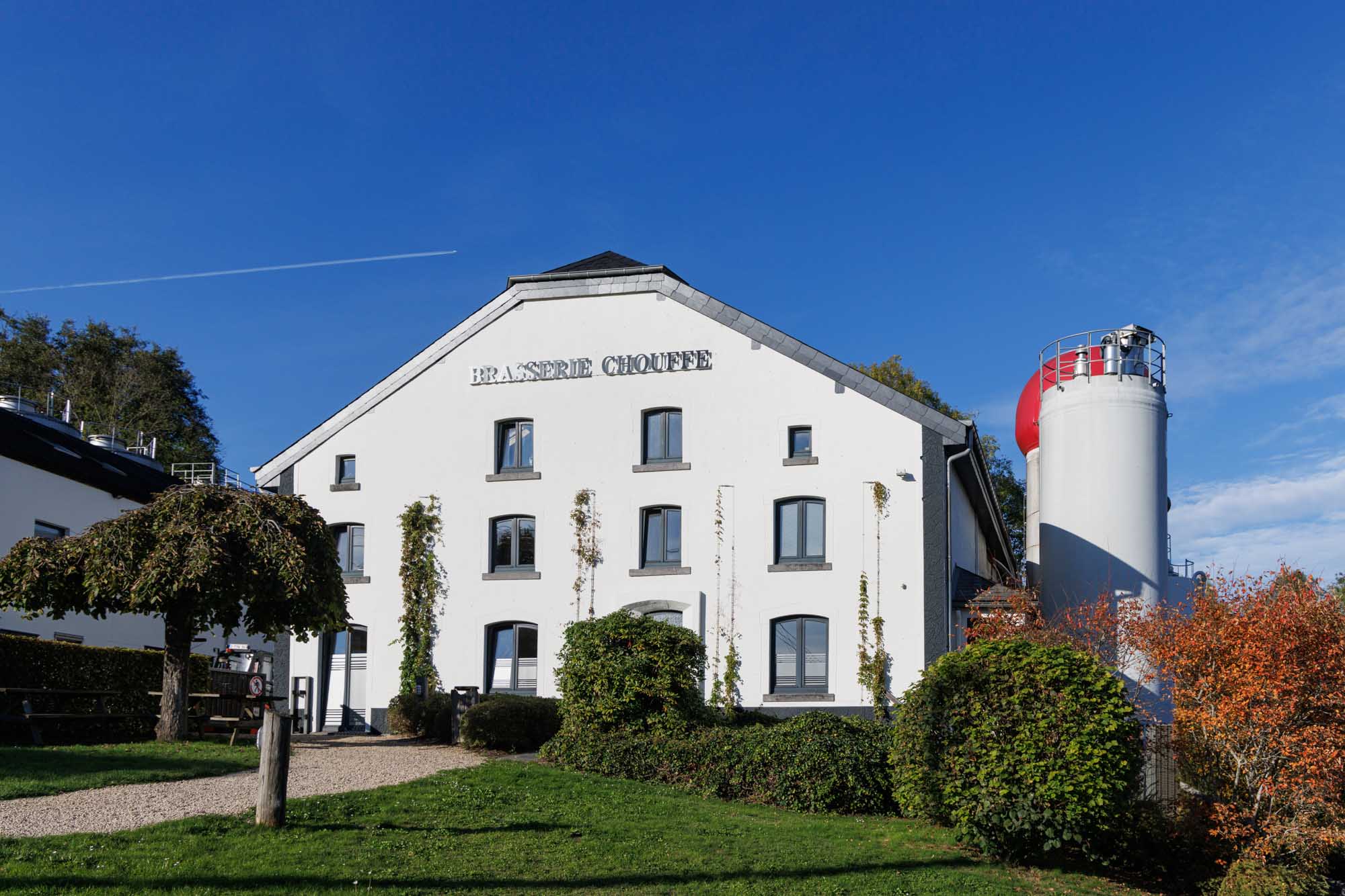 Façade de la Brasserie Chouffe à Achouffe avec son bâtiment blanc et ses installations brassicoles sous un ciel bleu.